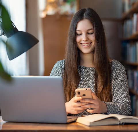 Young girl working at the table in university classroom. Female student reading books, prepare research as her home assignment using modern technologies: laptop and mobile phone with the Internet